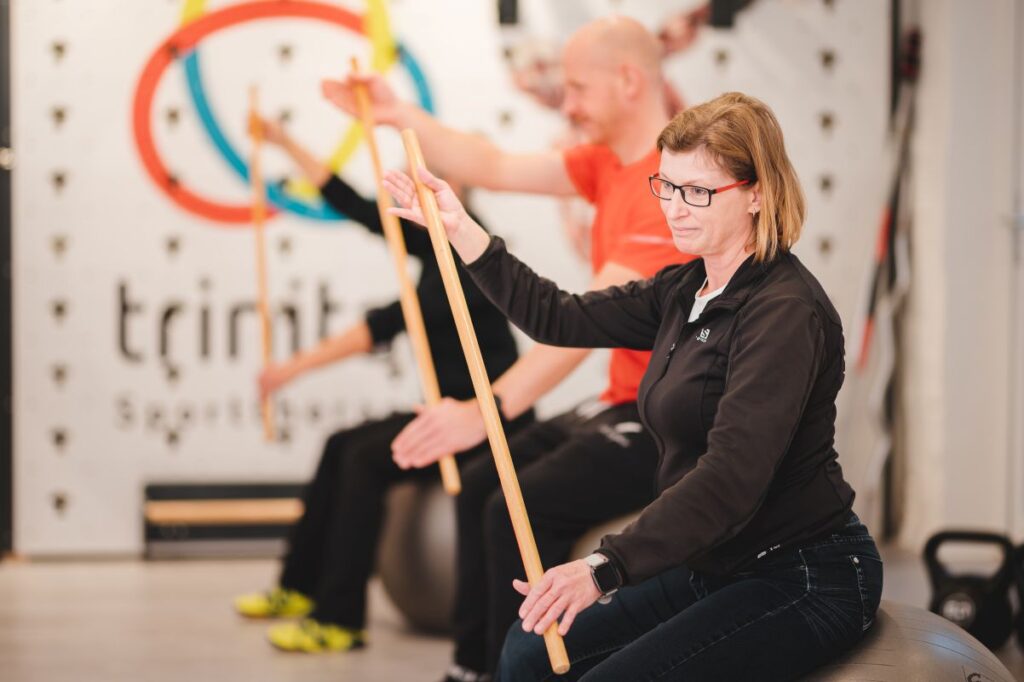 Bild von drei Personen sitzend auf einem Pezziball mit Gymnastikstab in der Hand. Im Hintergrund ist ein trinitas-Logo zu sehen.