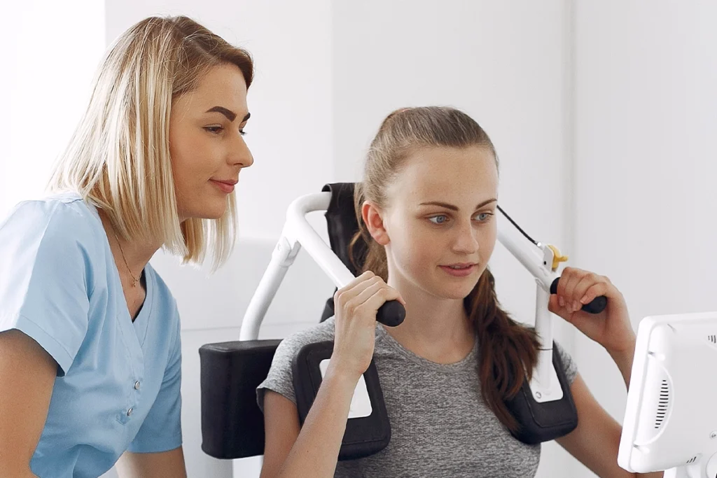Physiotherapeutin zeigt ihrer Patientin Übungen für den Schulterbereich mit einem Sportgerät während der Krankengymnastik am Gerät.